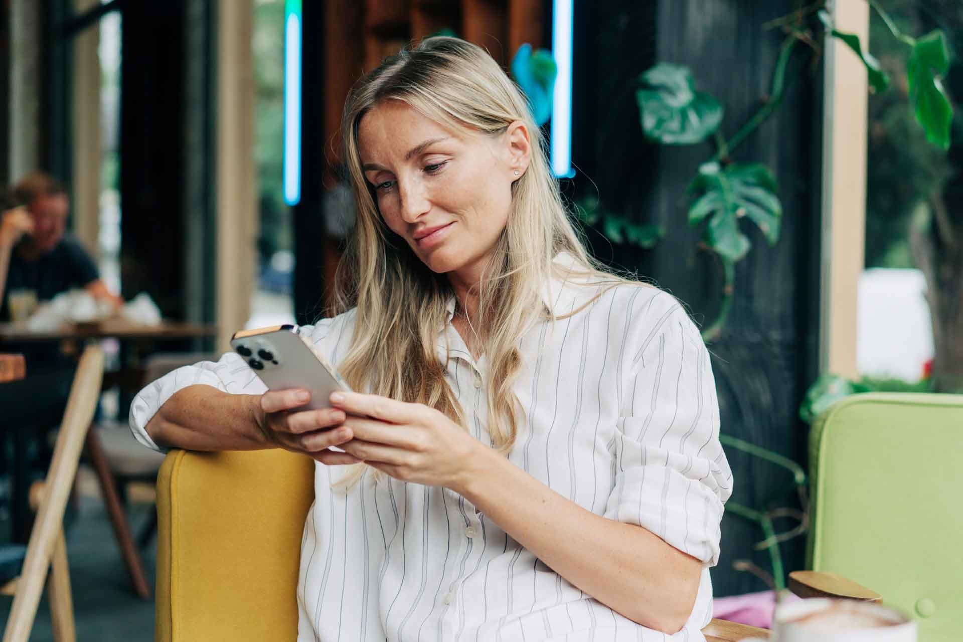Blonde Frau in gestreifter Bluse liest lächelnd auf ihrem Smartphone in einem Café