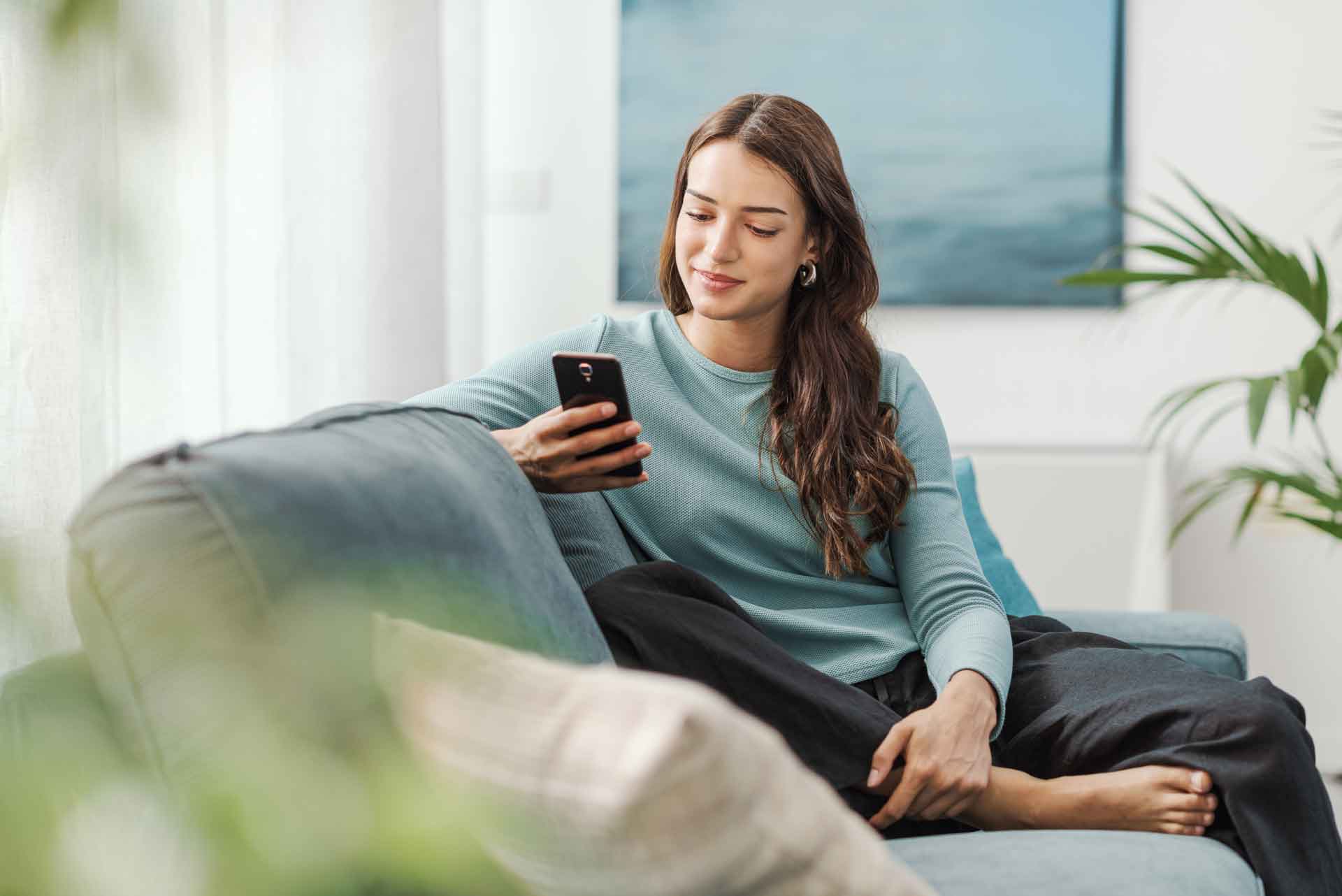 Frau mit langem braunem Haar liest entspannt auf dem Sofa auf ihrem Smartphone